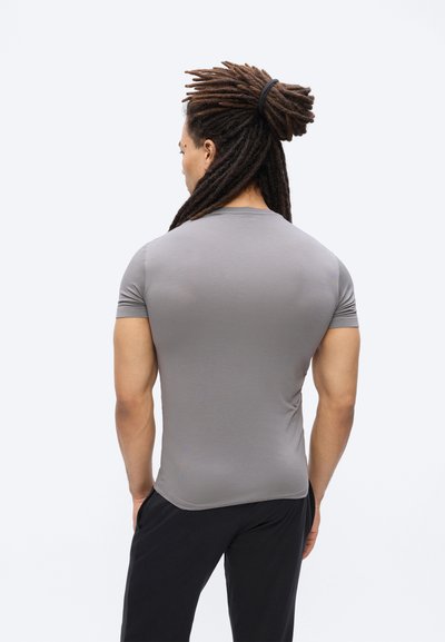 Άνδρας με καστανά dreadlocks δεμένα πίσω, φορώντας εφαρμοστό γκρι κοντομάνικο πουκάμισο και μαύρο παντελόνι, φωτογραφημένος από πίσω πάνω σε απλό φόντο.