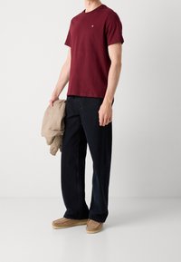 Burgundská bavlněná trička s krátkým rukávem a kulatým výstřihem, spárovaná s tmavými širokými kalhotami a béžovou bomber bundou, s muškově hnědou obuví.