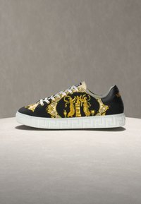 Black sneaker featuring gold baroque designs, white textured sole with Greek key pattern, and gray accents near the collar.