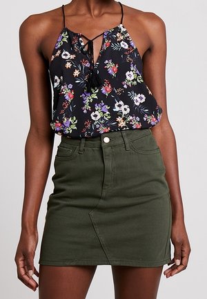 Eine Frau trägt ein schwarzes, geblümtes Spaghettiträgertop kombiniert mit einem olivgrünen, hoch taillierten Jeans-Minirock.