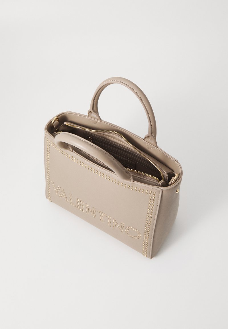 Sac à main en cuir beige avec des accessoires en or, deux poignées supérieures, un compartiment zippé et une accentuation de lettres cloutées à l'avant.