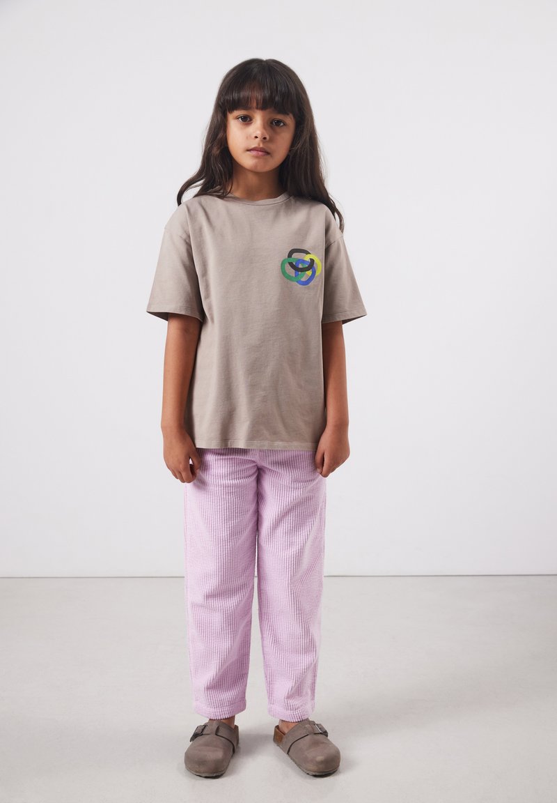 Junges Mädchen mit langen dunklen Haaren, das ein beiges T-Shirt mit einem mehrfarbigen Logo, hellrosa Hosen und braune Slipper trägt und drinnen steht.