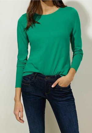 Frau trägt ein leuchtend grünes Langarmshirt und dunkelblaue Jeans, eine Hand in der Tasche, vor schlichtem Hintergrund.