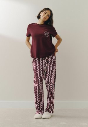 Woman in burgundy t-shirt with text and burgundy and pink wavy patterned pants standing against a plain light wall wearing white slippers.