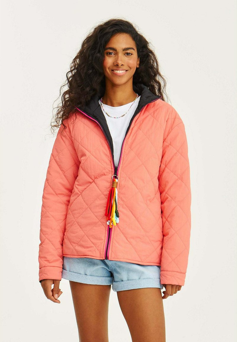 Prošívaná korálová bunda s černou kapucí, která má přední zip s vícebarevnými třásněmi a volný střih, doplněná džínovými šortkami.