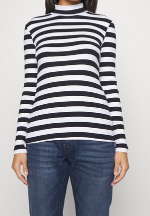 Kvinde iført en sort- og hvidstribet langærmet rullekravstrøje vandret og blå jeans med hænderne afslappet ned langs siderne.