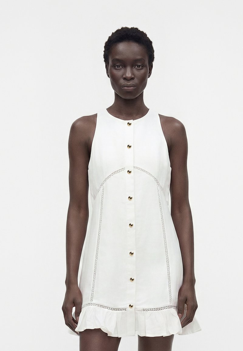 Femme debout portant une robe blanche sans manches avec boutons sur le devant, ourlet à volants et détails en dentelle sur fond uni.
