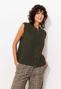 Débardeur en tricot vert olive sans manches avec une fermeture éclair à l'avant, présentant un motif texturé. Associé à un pantalon à carreaux beige et marron.