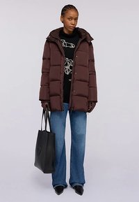 Hnědá péřová bunda s kapucí, krabicovitý střih a viditelné švy. Kombinována s černým svetr s potiskem, rozšířenými džínami a černou taškou přes rameno.