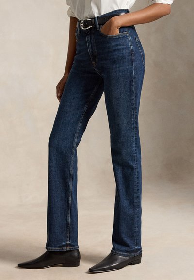 Persona che indossa jeans blu scuro a gamba dritta, cintura nera, camicia bianca infilata e stivali neri a punta, in piedi con una mano in tasca.