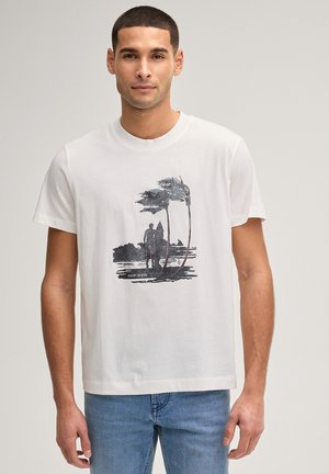 Mężczyzna w białej koszulce z czarnym nadrukiem osoby trzymającej deskę surfingową obok dwóch palm, w niebieskich dżinsach, na neutralnym tle.