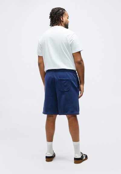 Άνδρας με πλεξούδες, που φοράει λευκό μπλουζάκι, σκούρο μπλε σόρτς με πίσω τσέπη, λευκές κάλτσες και ασπρόμαυρα αθλητικά παπούτσια, στέκεται γυρισμένος με την πλάτη.