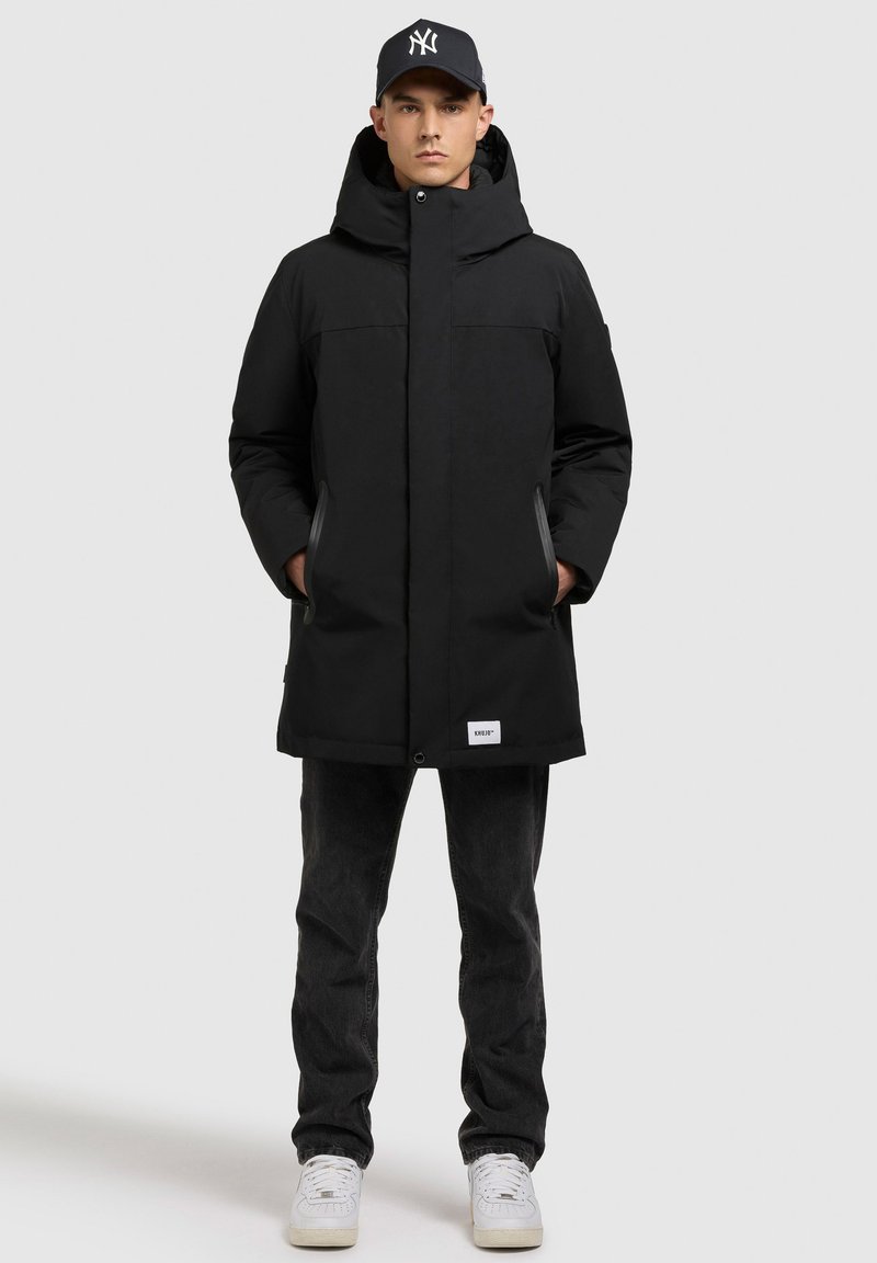 Hombre con un abrigo de invierno negro con capucha, gorra de béisbol negra, jeans negros y zapatillas blancas, de pie con las manos en los bolsillos del abrigo.