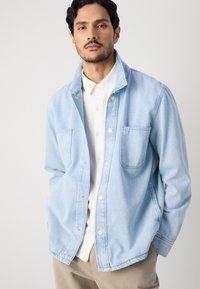 Light blue denim jacket with a collar, snap buttons, and two front pockets, layered over a white button-up shirt, structured fit.