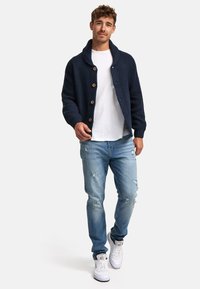 Cárdigan de punto azul marino con grandes botones de madera, llevado sobre una camiseta blanca y unos vaqueros claros desgastados, combinado con zapatillas blancas.