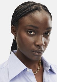 Himmelblaues Hemd mit Kragen und Knopfdetails, kombiniert mit einer goldenen Kettenhalskette. Die Haare sind zu Zöpfen gestylt, mit einem glatten Teint.