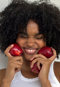 Punaiset omenat, kiiltävä kuori, pidetään molemmissa käsissä; henkilö, jolla on kihara musta hius, hymyilee, päällä valkoinen toppi vaaleaa taustaa vasten.