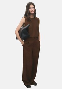 Bruine mouwloze crop top gecombineerd met hoge taille bruine wijde broek. Model houdt een zwarte gestructureerde handtas vast. Soepele stof.