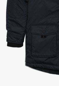 Manga y sección inferior de una chaqueta acolchada azul marino con puño acanalado y bolsillo con solapa asegurado por un botón de presión negro y una etiqueta negra con costura naranja.