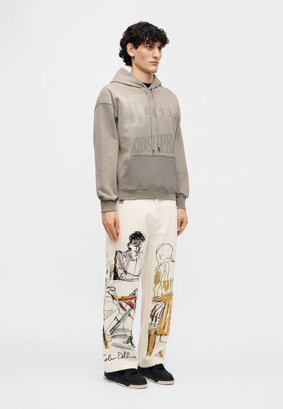 Γκρι φούτερ με κουκούλα και μπροστινή τσέπη, με τυπωμένο κείμενο, σε συνδυασμό με λευκό παντελόνι που έχει πολύχρωμα καλλιτεχνικά σκίτσα και μαύρα αθλητικά παπούτσια.