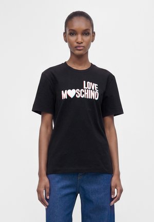 Γυναίκα που φοράει μαύρο T-shirt με "LOVE MOSCHINO" και σύμβολο καρδιάς, συνδυασμένο με μπλε τζιν, στέκεται μπροστά σε ένα απλό φωτεινό φόντο.