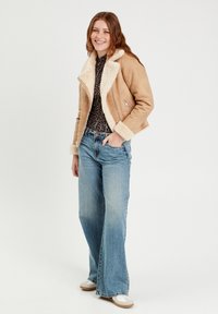 Chaqueta de ante marrón con cuello y puños de shearling blanco, detalle de cremallera plateada, combinada con vaqueros acampanados de color azul claro y zapatillas blancas.