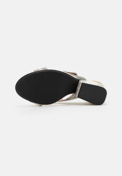 Sandales à talons avec un dessus texturé argent et noir, une semelle en caoutchouc noir et des brides décoratives. Le talon est de forme bloc pour plus de stabilité.