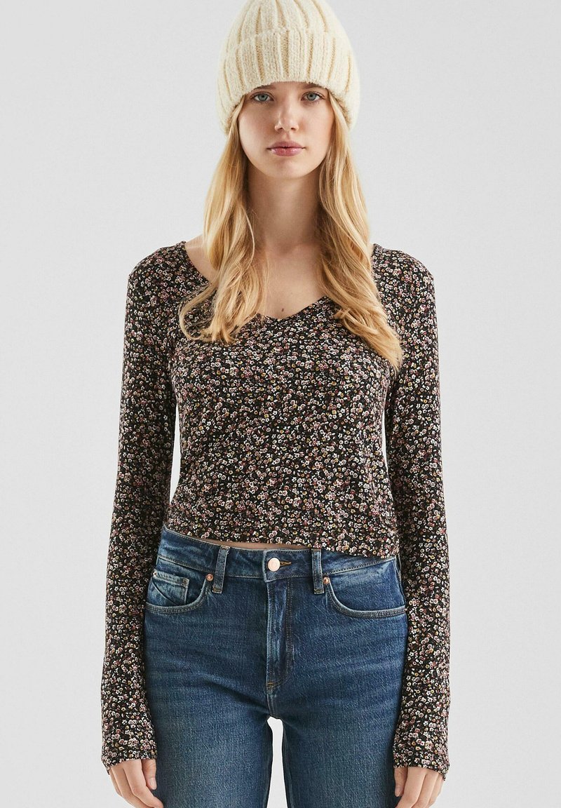Langarm bloemen blouse met een diepe V-hals, zwarte achtergrond en patronen van kleine roze, witte en gele bloemen; gecombineerd met blauwe jeans.