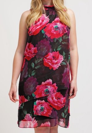 Czarna sukienka bez rękawów z warstwową przezroczystą tkaniną i dużym, jasnoróżowo-czerwonym kwiatowym wzorem, noszona przez stojącą kobietę.