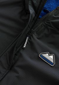 Črna jakna z zadrgo, ki se ponaša z teksturirano zunanjostjo, modrim flis podlogom in logotipom z geometrijskimi vzorci ter blagovno znamko.