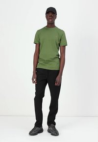 T-shirt de algodão verde com mangas curtas, combinado com calças pretas e ténis cinzentos. O t-shirt apresenta um pequeno logótipo no peito.