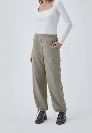 Person trägt ein weißes, langärmliges, eng anliegendes Oberteil, lockere olivgrüne Cargohosen mit elastischem Bund an den Knöcheln und schwarze Schnürschuhe, steht mit der Hand in der Tasche.