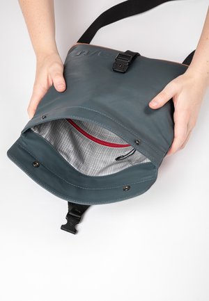 Hände halten eine dunkelgraue Umhängetasche offen, mit einer inneren Tasche mit rotem Reißverschluss und einem Innenfutter mit Rastermuster.