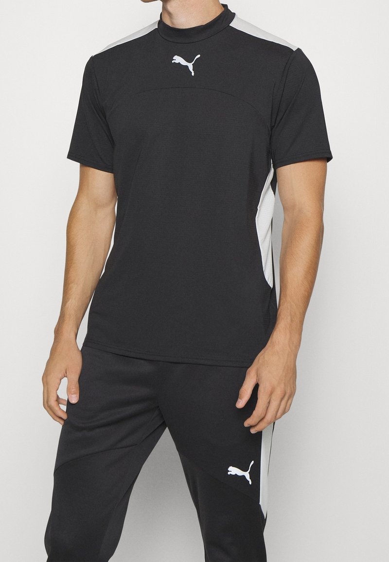 Hombre vestido con una camiseta deportiva negra de Puma y pantalones de chándal a juego, con logo blanco y paneles laterales, de pie frente a un fondo blanco.