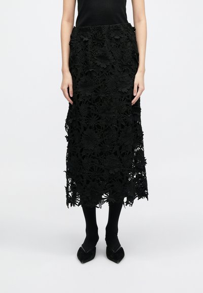 Fekete csipke szoknya bonyolult virágmintákkal, középhosszú kivitelben, textúrált felülettel és fodros szegéllyel, fekete harisnyával és cipővel párosítva.