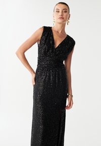 Sort selskabskjole med palietter, dyb V-udskæring, kortærmer og en indsnævret talje. Har en sideslids og en struktureret, glitrende overflade.