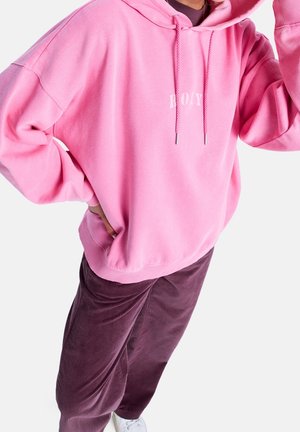 Personne portant un sweat à capuche rose ample avec un texte blanc, un pantalon en velours côtelé bordeaux et des baskets blanches, posant avec une main sur la hanche.