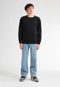 Sudadera negra con cuello redondo y pequeño logo en forma de círculo; combinada con jeans azul claros y zapatillas en blanco y negro. Textura suave, ajuste casual.