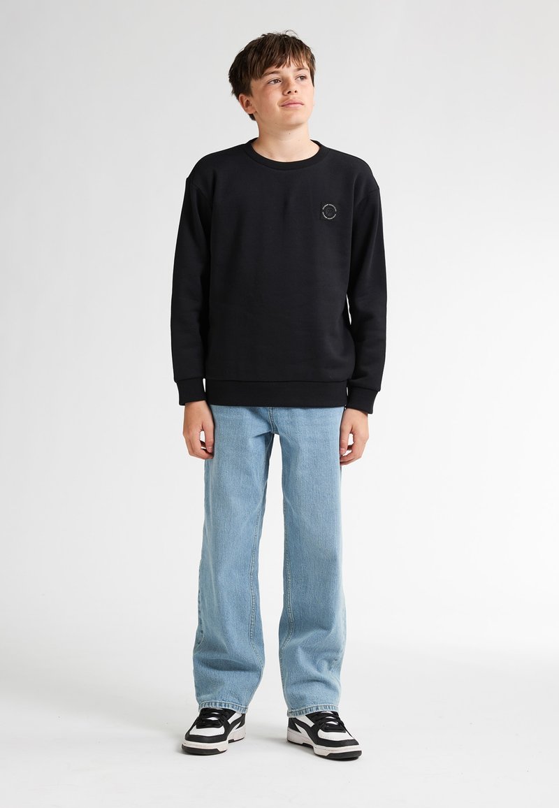 Sudadera negra con cuello redondo y pequeño logo en forma de círculo; combinada con jeans azul claros y zapatillas en blanco y negro. Textura suave, ajuste casual.