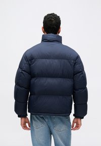 Σακάκι τύπου puffer σε ναυτικό μπλε με ψηλό γιακά, με επένδυση και ελαστικούς καρπούς. Περιέχει λογότυπο στην πλάτη. Συνδυάζεται με ανοιχτό μπλε τζιν.