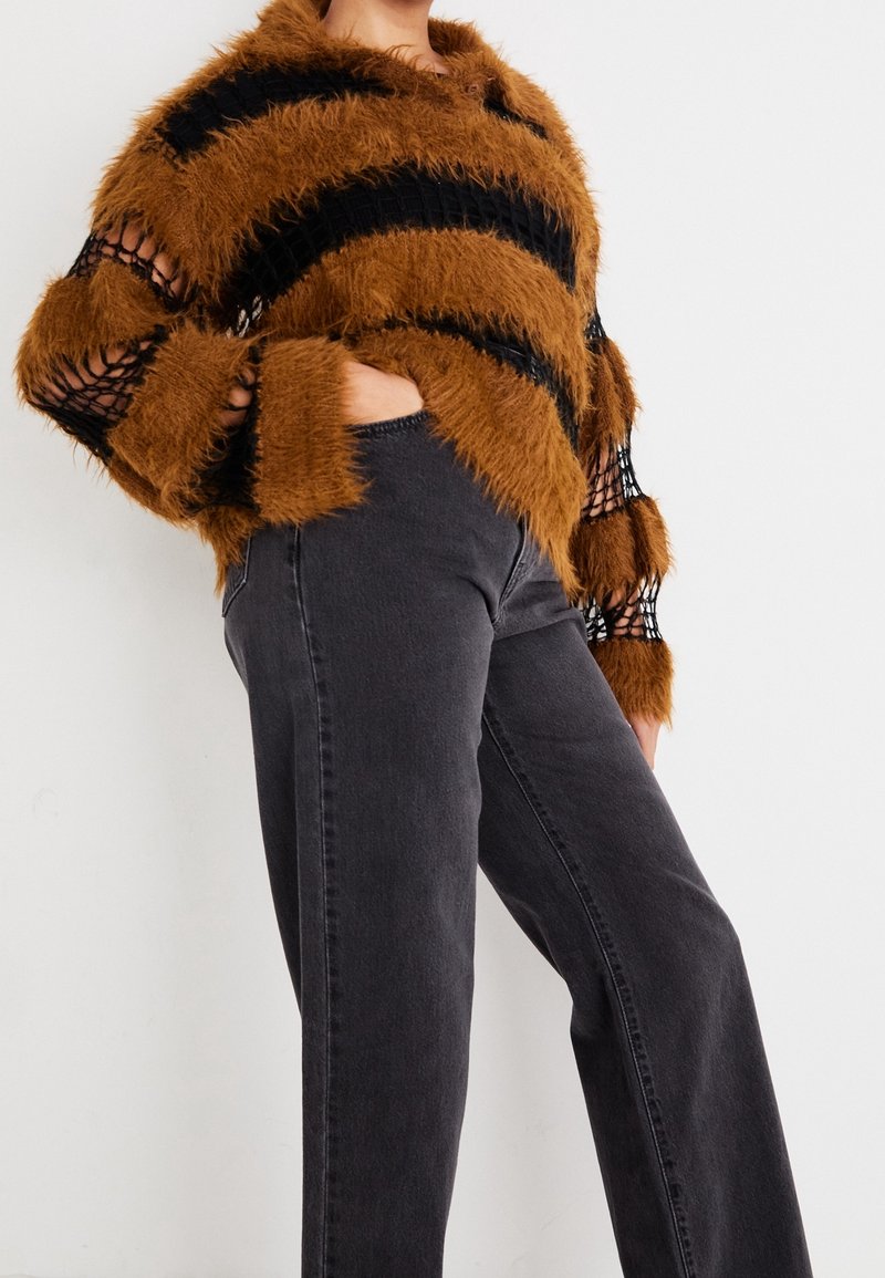 Personne portant un pull pelucheux rayé marron et noir avec des manches en résille et un jean taille haute gris foncé, une main dans la poche.