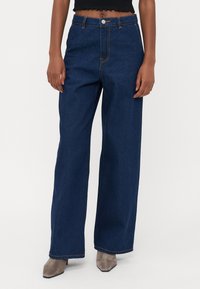 Ballonjeans aus blauem Denim mit hochgeschnittenem Bund, versehen mit gelben Nähten und einem Knopfverschluss. Glatte Textur mit klassischem Design.
