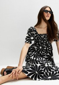 Zwart-wit bloemenjurk met korte puffmouwen, een vierkante halslijn en een gesmokt lijfje. Model draagt een zwart zonnebril en platform sandalen.