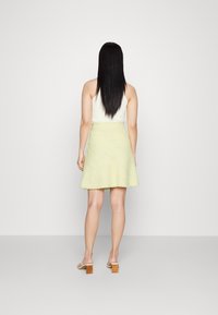 Rok anyaman kuning muda dengan desain melebar, pola tekstur, dan hem mentah. Dipadukan dengan atasan tanpa lengan putih dan sandal bertumit blok.