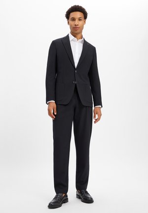 Man standing wearing a black suit, white dress shirt, and black dress shoes against a plain white background.
