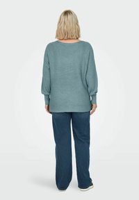 Una mujer rubia está de espaldas, vistiendo un suéter holgado de canalé color turquesa con mangas abullonadas y unos jeans anchos de color azul oscuro.