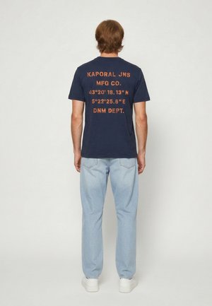 Homme aux cheveux châtain clair portant un t-shirt bleu marine avec des coordonnées imprimées et un jean bleu clair, debout devant un fond uni.