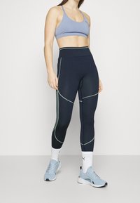Leggings blu navy con cuciture verdi a contrasto, design a vita alta, abbinati a un reggiseno sportivo azzurro e scarpe da ginnastica azzurre.