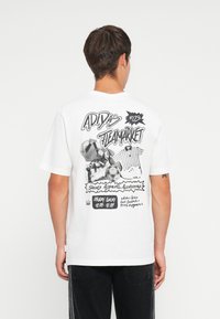 Biele tričko s grafickým dizajnom na zadnej strane, obsahujúcim čierny text a ilustrácie týkajúce sa značky Adidas, s voľným strihom a krátkymi rukávmi.