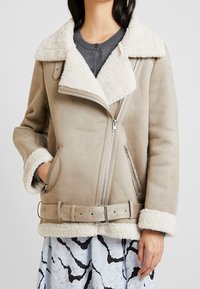 Beige shearling-lined jacket with a zip closure, wide lapels, and adjustable belt. Features side pockets and contrasting textured cuffs.
